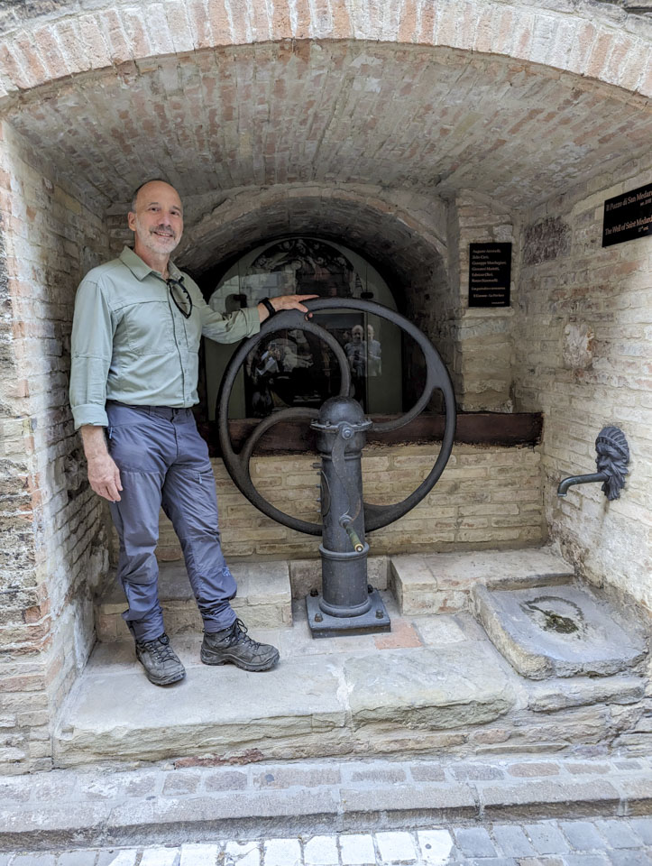 Paul next to a black wheel with a handle, with a water spigot on the wall opposite him