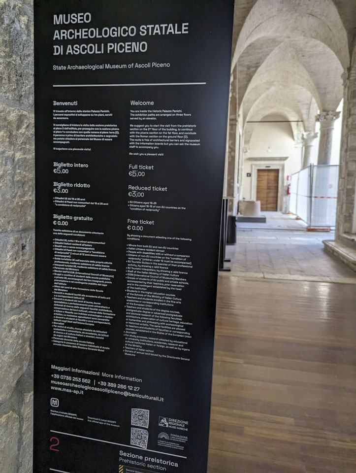 Entrance sign to the Ascoli Piceno archeological Museum, with a long hallway behind it and a large wooden door at the end of the hall