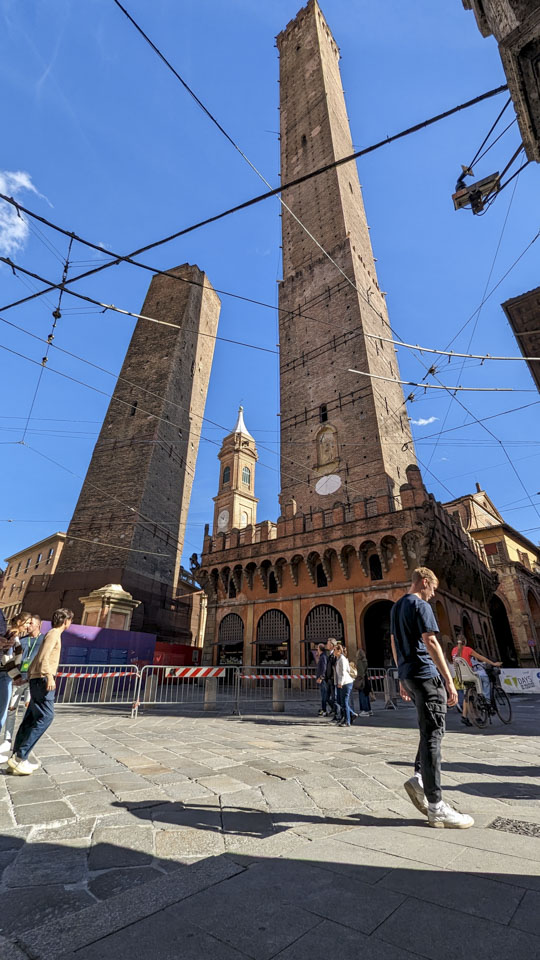 A photo of the Two Towers of Bologna taken from near the base, looking up. Some wires obscure the view.