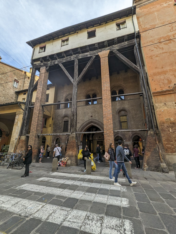 A building with one story that sticked out over the sidewalk and very tall supports, some wood and some brick, reaching down to the ground.