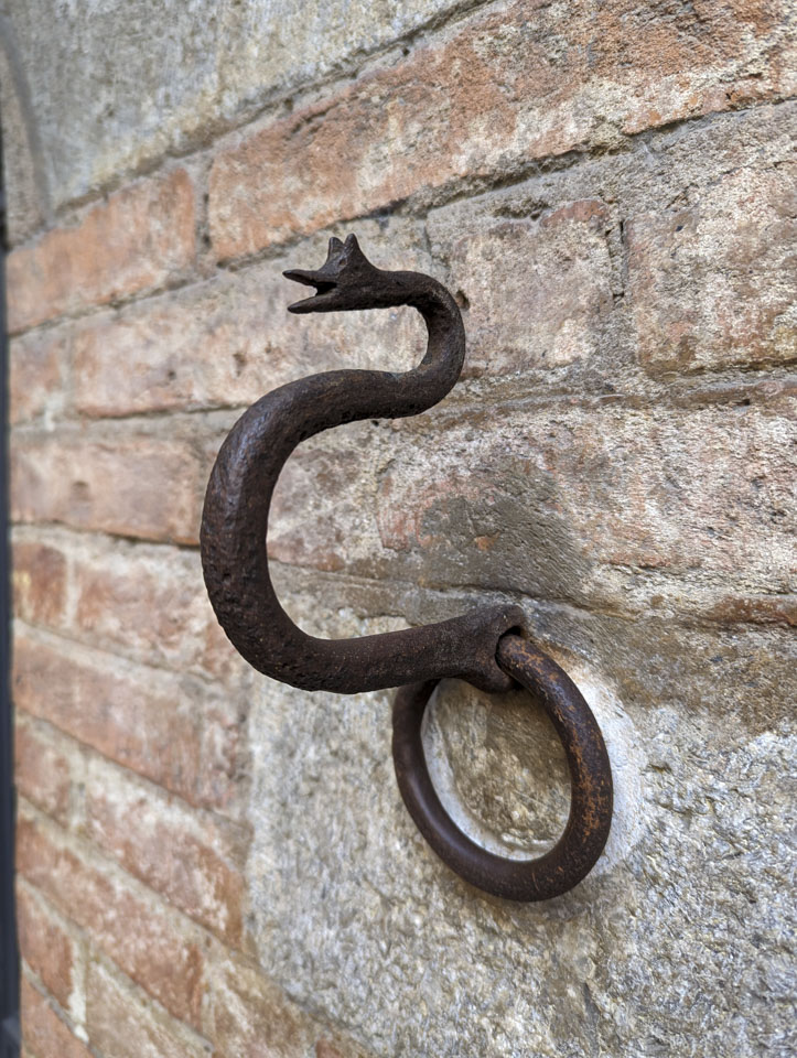 A small metal decoration on the side of the building, with a face at the top and a curved loop. 