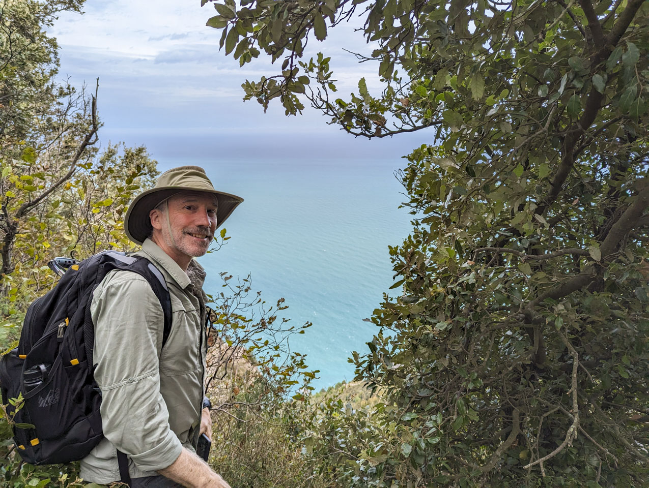 Paul is looking towards the camera, with a hat and backpack on. There are trees and shrubs on each side of the photo with a small clear area in the middle through which the sea can be seen.