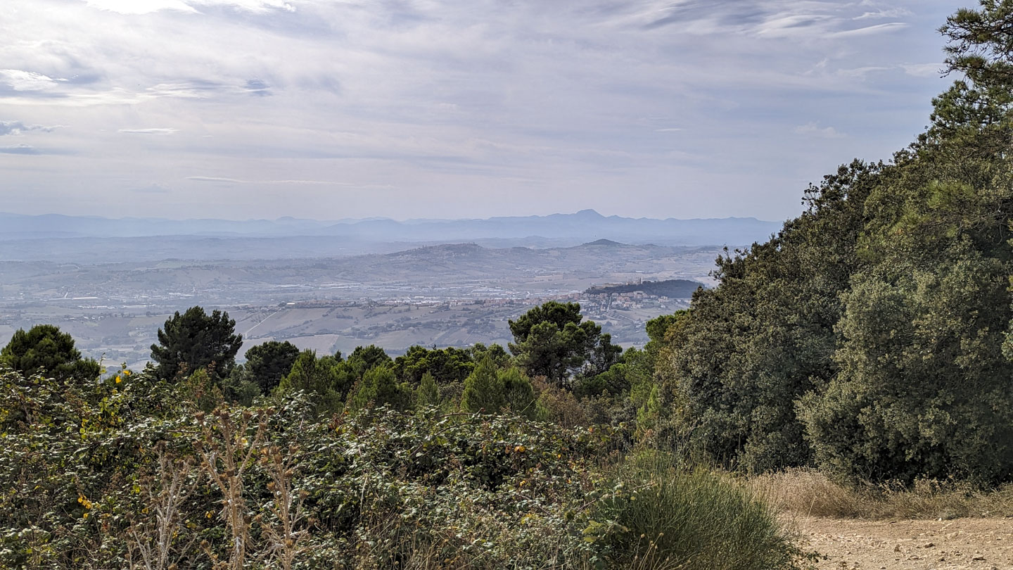 A cloudy day with mountains spread across the horizon. Closer are some hills, then agricultural land. In the foreground are shrubs and a dirt path.