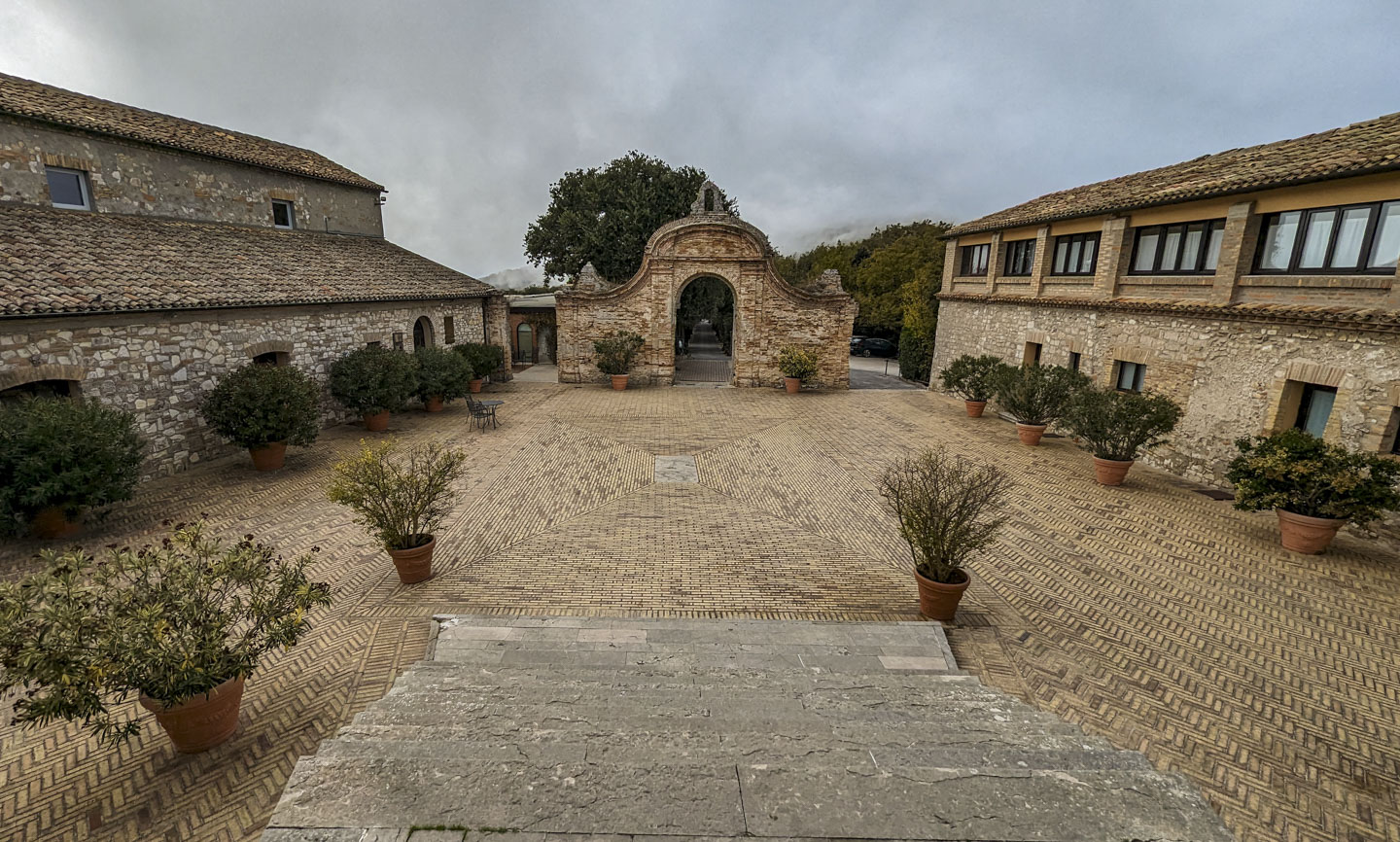 Looking down a pale stone staircase, across a stone courtyard, to a partial wall with an arched entrance. On each side of the courtyard are stone buildings, and there are many potted plants in the courtyard. Some fog can be seen in the distance.