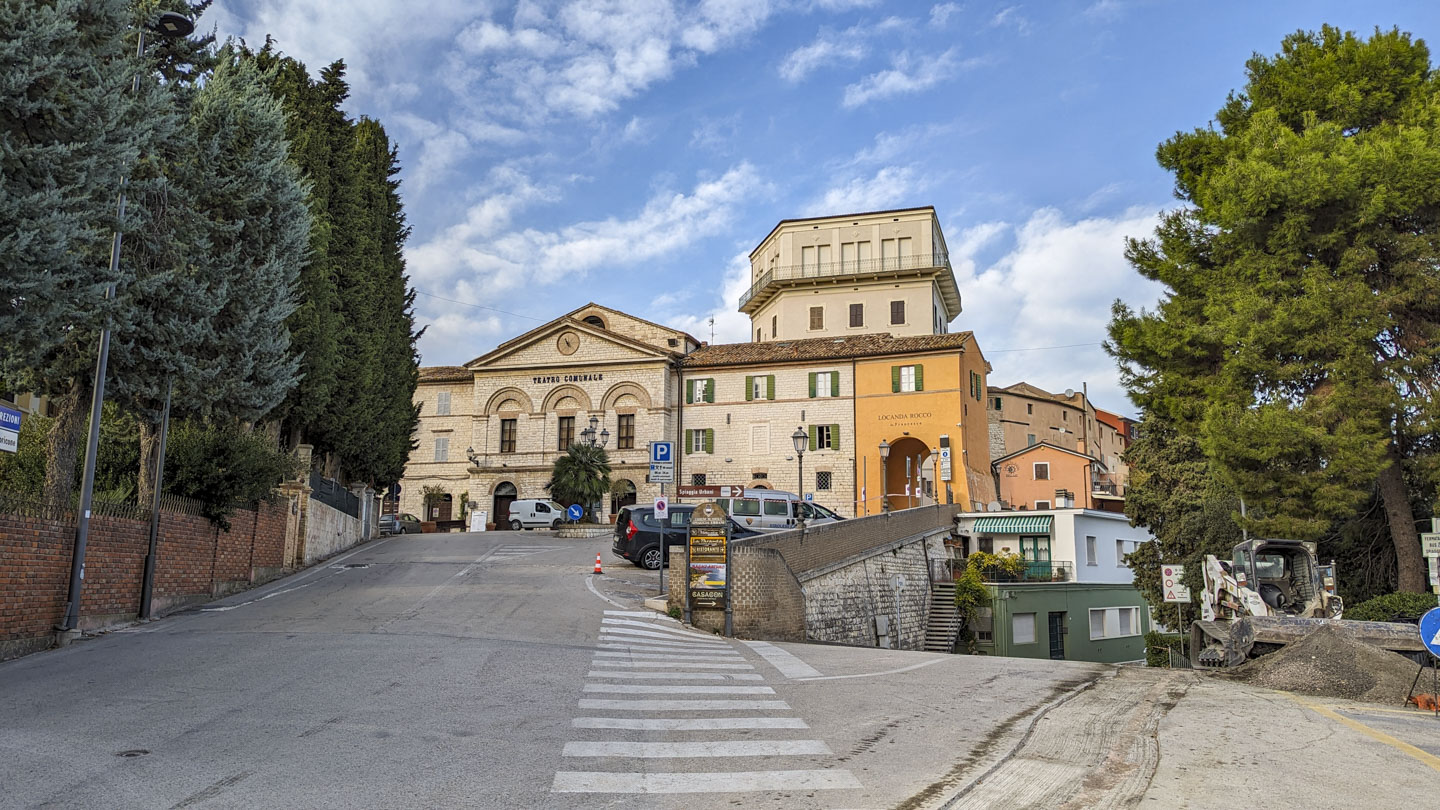The viewer is standing at a split in the road. To the right is a steep road down, and straight goes up to old stone buildings and a city theater building.