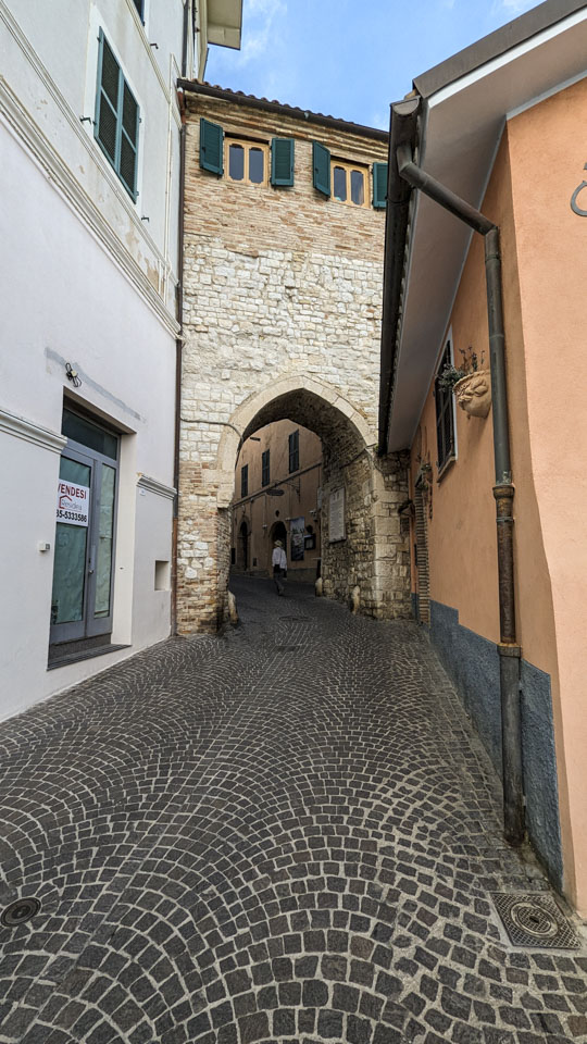 Anne can be seen in the distance going under what is likely the original city wall after having walked up a stone road. There is a store front on the left that is for sale.