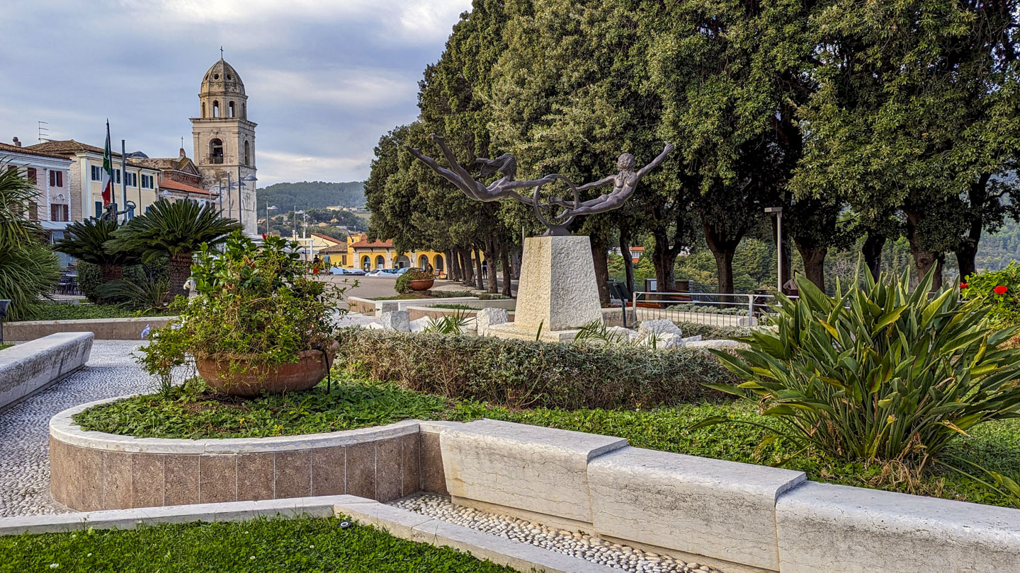 Standing in a small park with plants and paths, you are looking over it towards a plazza.  Tall trees are on the right and buildings are on the left.