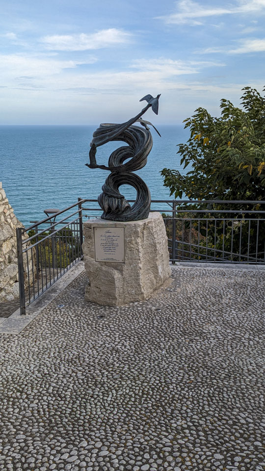 A sculpture that seems to represent birds on swirls of wind. Behind it is the Adriatic sea.