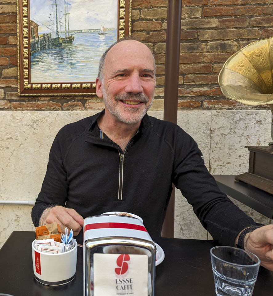 Paul sitting at an outdoor table in Fano. There is a water view painting on the wall behind him.
