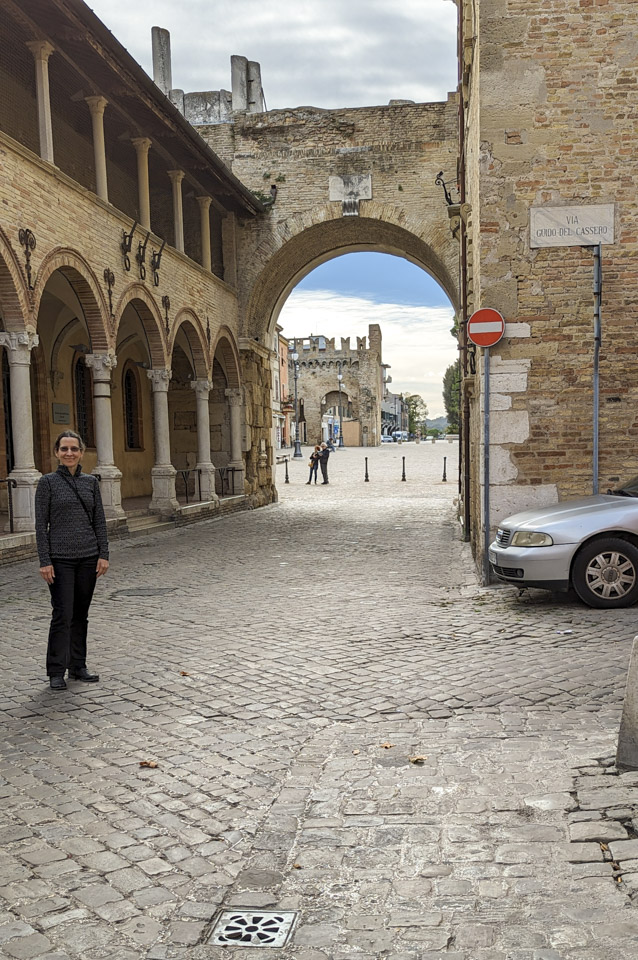 Anne is standing on the left in front of a line of stone archways, and in the center of the photo is a large archway. Through the arch can be seen the remains of an old city wall.