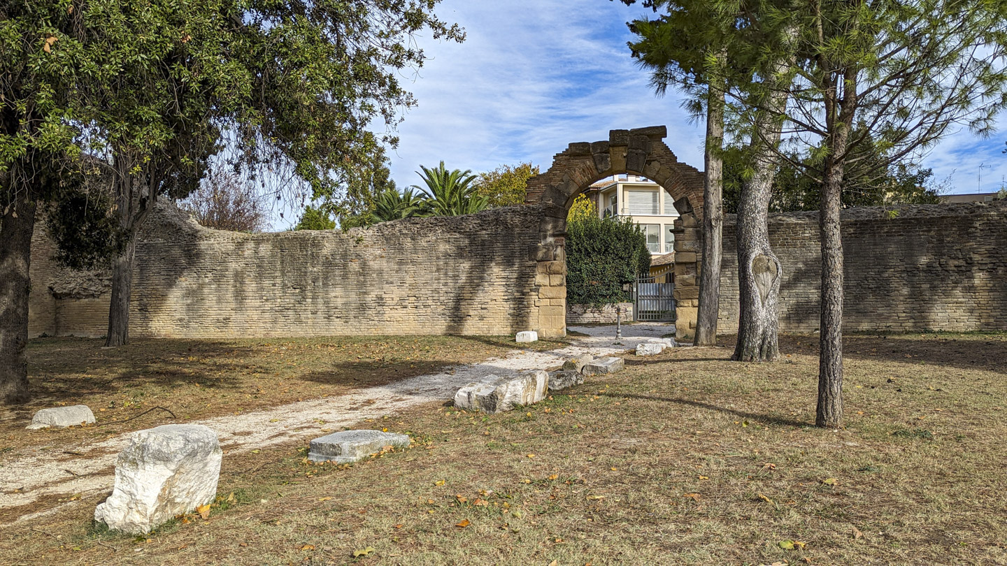 A gate in the wall to Fano, with a path leading up to it. There are some large stones on the side of the path and some tall trees can be seen. 