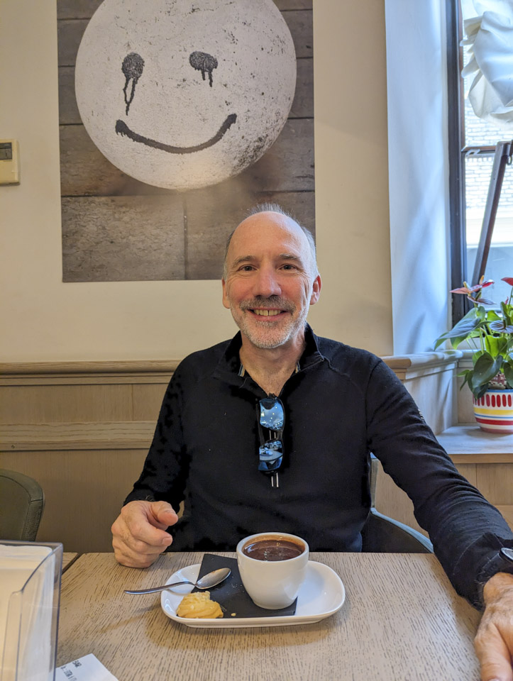 Paul sitting at a table in Caffè Centrale in Fano, with a hot chocolate in front of him. On the wall behind him is a print of a what looks like a ping pong ball with a smiley face painted on it in runny ink.
