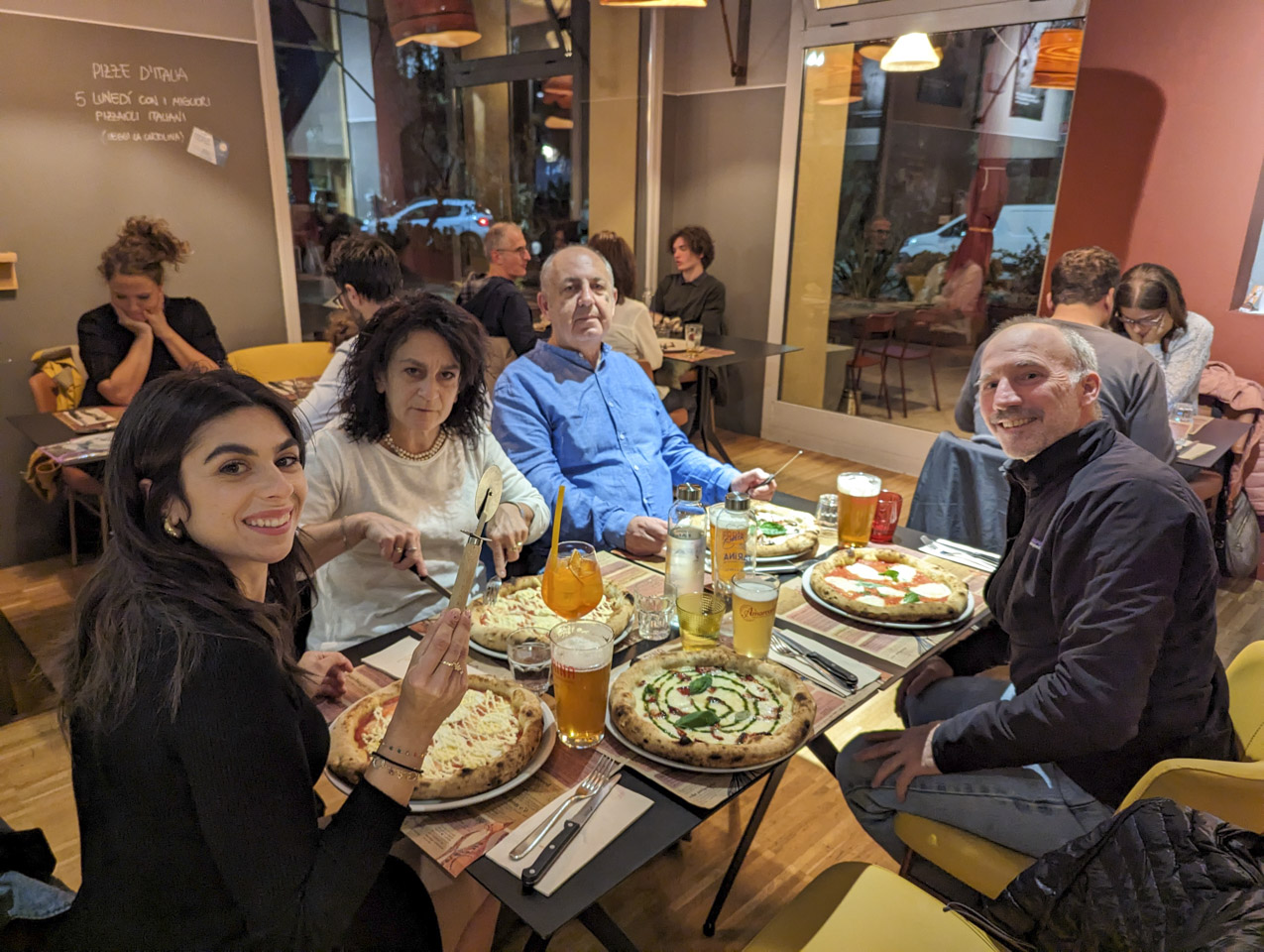 4 people sitting at a table with pizzas in front of each, plus one in front of an empty seat.