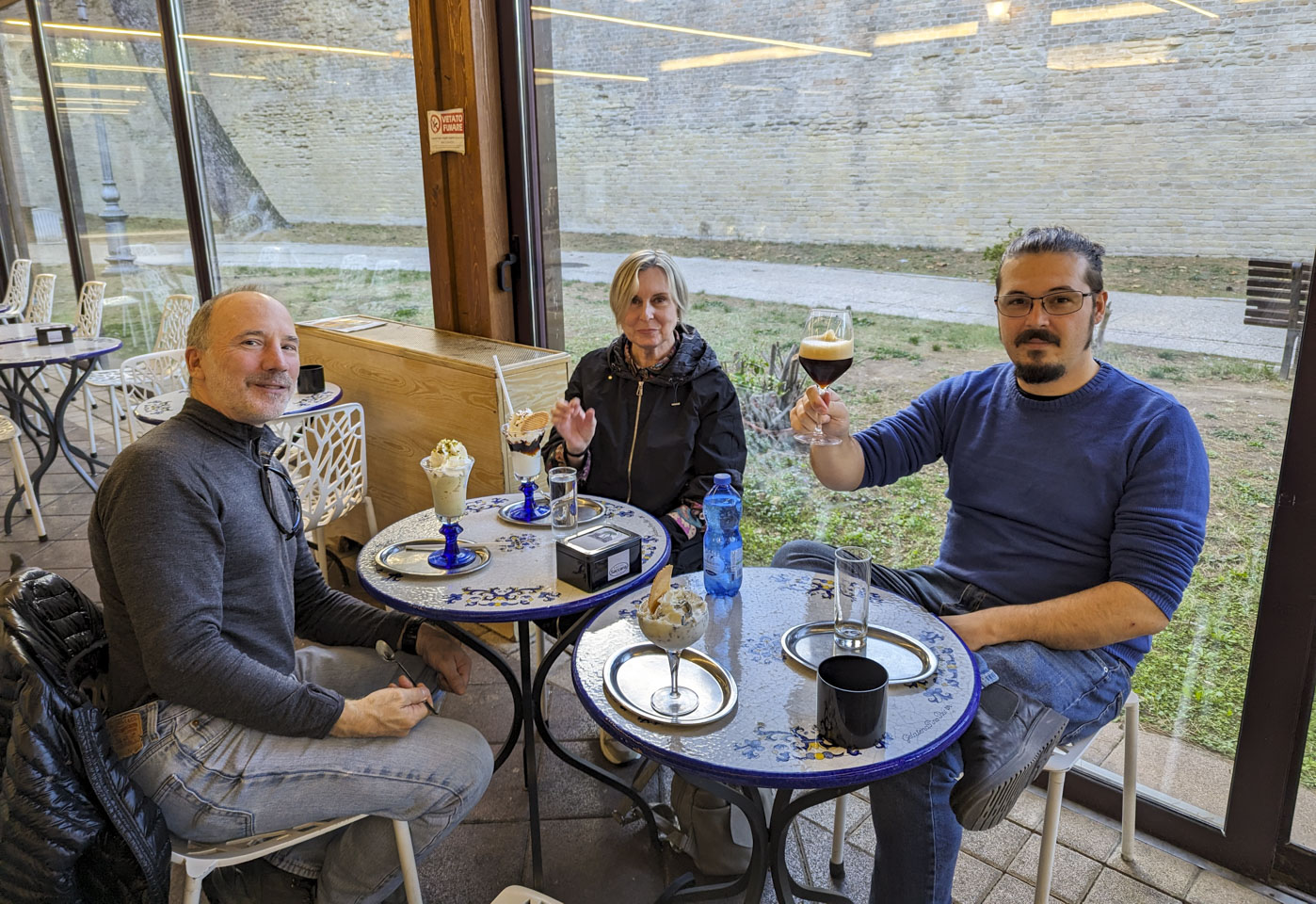 Paul, Giovanna, and Filippo sitting at two small round tables with some ice cream in fancy glasses. Filippo is drinking a beer instead of having ice cream.