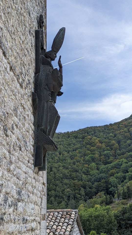statue of a saint on the Fonte Avellana exterior wall, with the forested hillside visible behind it