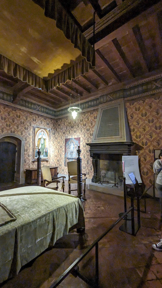 A room with a bed, fireplace, a couple of sitting chairs, and paintings on the walls.
