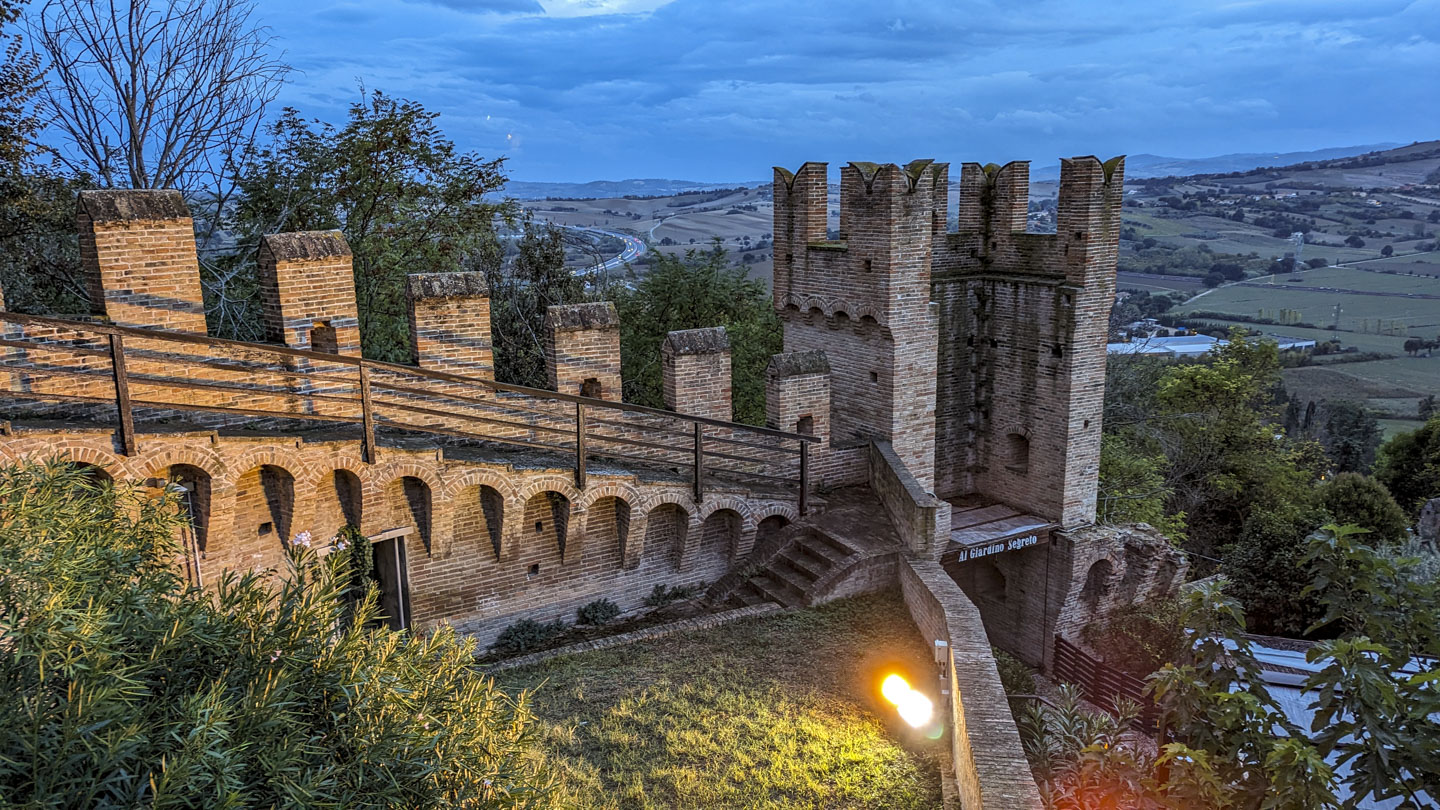 An evening view from a high point, with a descending castle wall leading to a tower with only 3 sides. Beyond that can be seen a highway, farmland, and a few mountains in the far distance.
