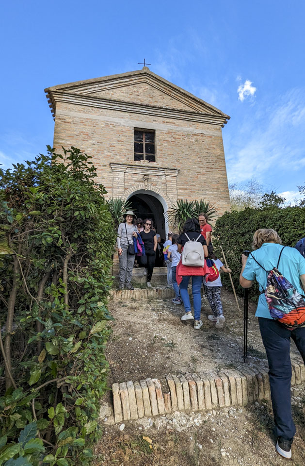 Anne and others outside of a tall but narrow stone chapel with a dirt walkway up to it.