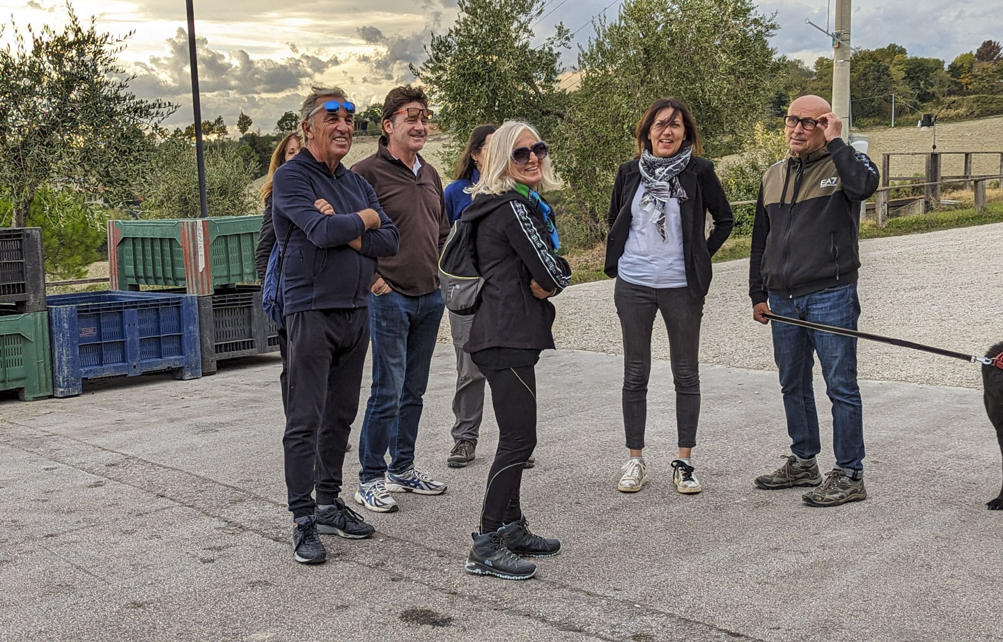 People standing on a paved area with some containers and olive trees in the background.