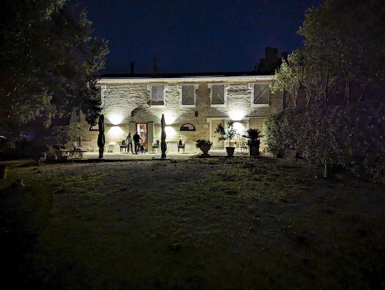 A two story long stone house with lights on the exterior of the house. Two men and a dog are standing in front of an open door into the house. There are some chairs stacked on the patio, and trees on each side of the yard.