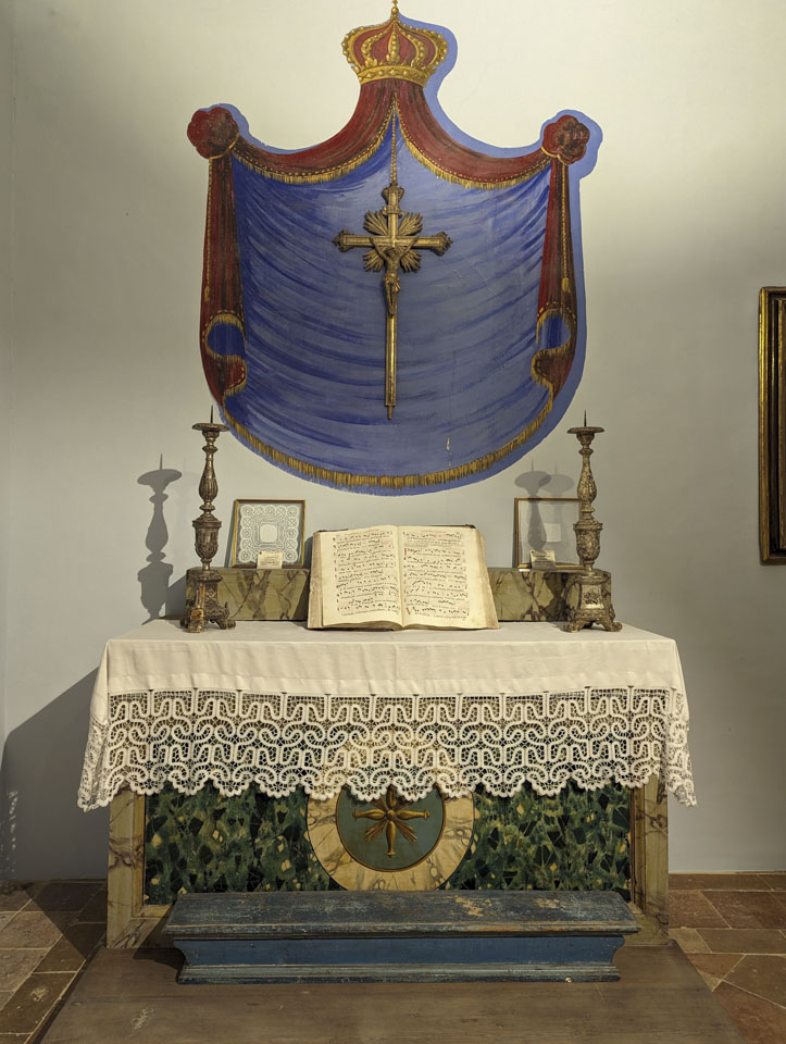 A religious altar with a lace trimmed altar cloth. On the wall above is a painted blue shield and a cross.