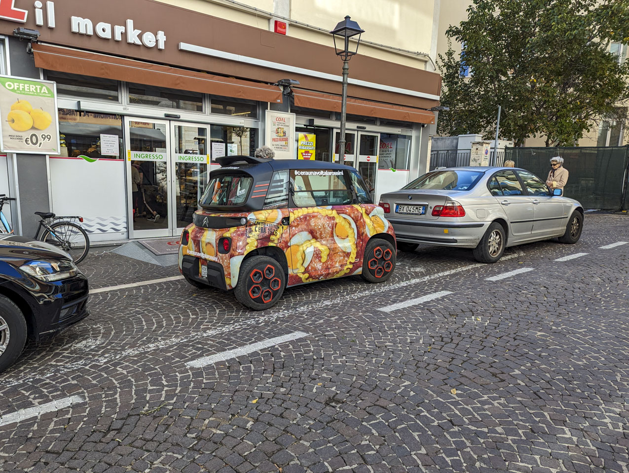 A photo of a street in front of a market with a few vehicles. One is a small vehicle with a black top and the sides custom painted with the image of a Rossini pizza, which has hard boiled eggs and mayonnaise on it.