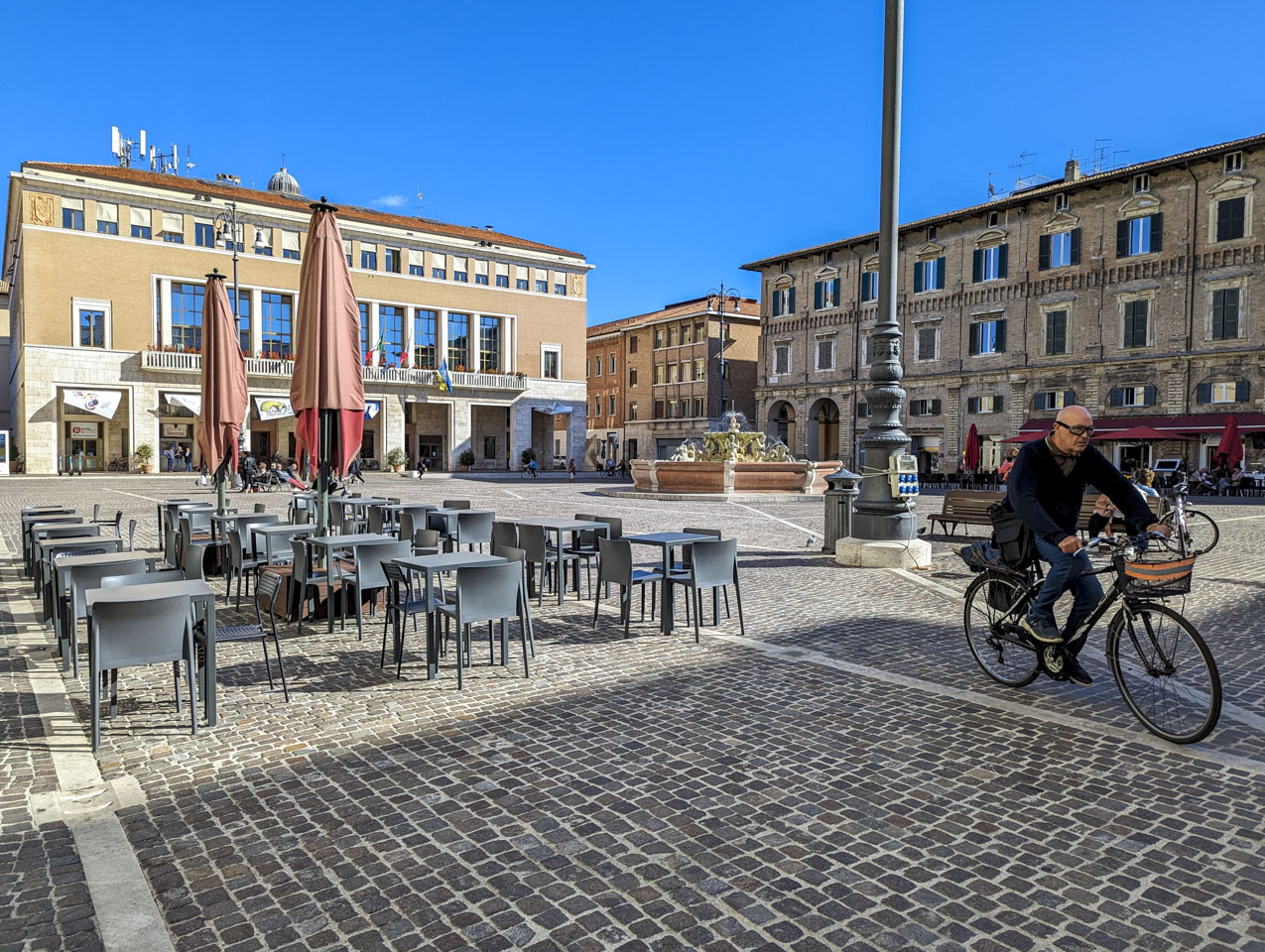 A plaza with a stone pavement, a fountain, and large buildings along the sides. A man is riding a bicycle and some people are sitting on some benches or chairs.