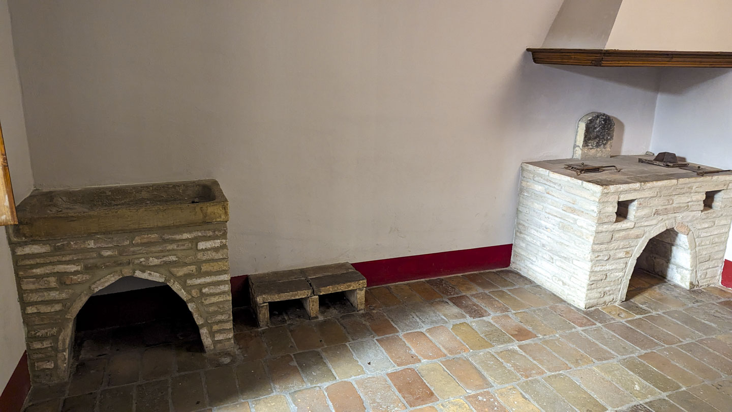 A room with a brick floor, On the right is a stone stove with a flat top and an exhaust chimney above it. On the left is a basic.