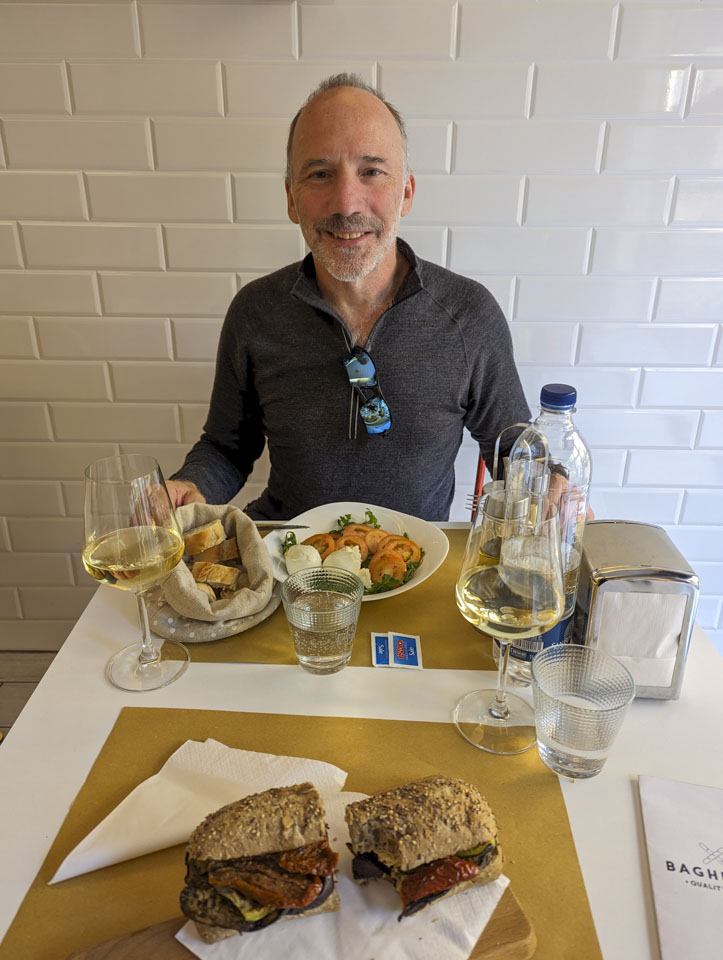 Paul is sitting in front of a white brick wall, behind a table.  On the table is a sandwich with eggplant and sundried tomatoes visible, a plate with tomato, mozzarella, and greens, two glasses with some white wine, a bottle of water, and some bread in a cloth bag.