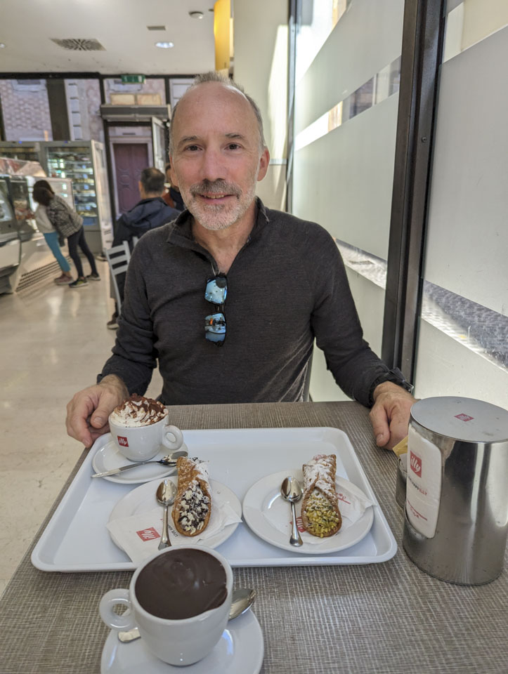 Paul is sitting at table with a tray in front of him. On the tray is a cup with whipped cream, and 2 cannoli. Closer to the camera is a cup of thick hot chocolate with no whipped cream.