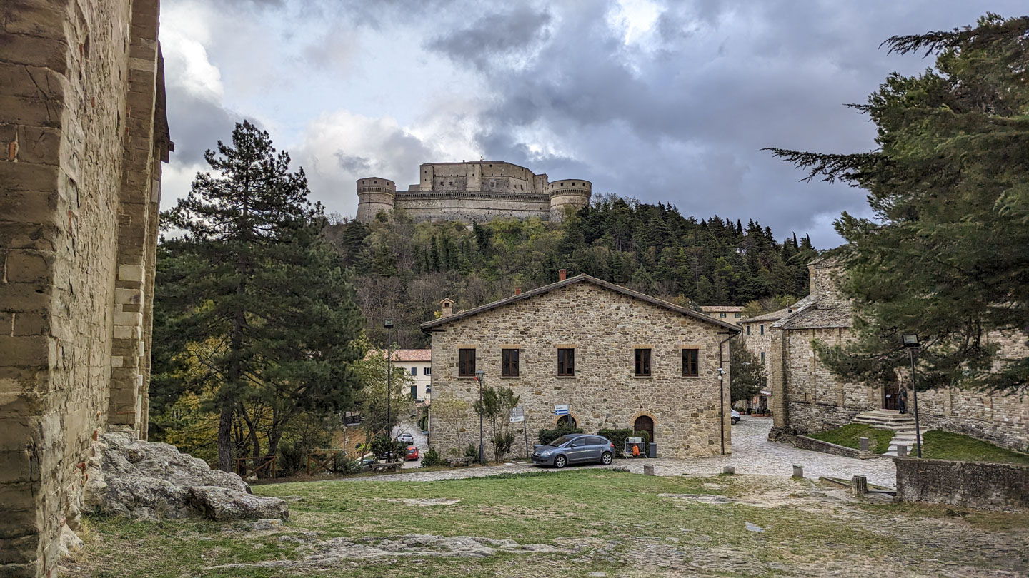 The stone wall of the Cathedral is on the left, the stone Parish Church is on the right, and there is a small stone building in the center. Behind this, high on a hill, is a very large stone fort castle with brooding clouds in the sky.