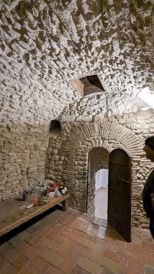 A small stone room with Roberto partially seen on the right. A small door leads to a very steep set of stairs. There is a low wooden platform on the left with a few artificial flowers on it. In the ceiling is a square wooden opening.