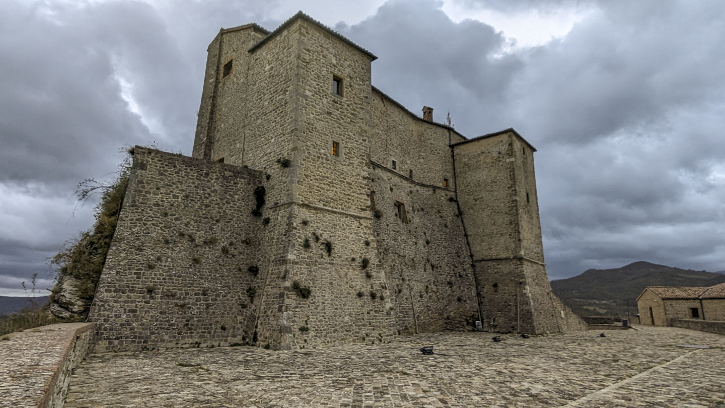 The stone fortress, perhaps 6 stories tall, has a couple of square towers protruding from the side towards us. A mountain can be seen in the distance. The sky is cloudy and gray.