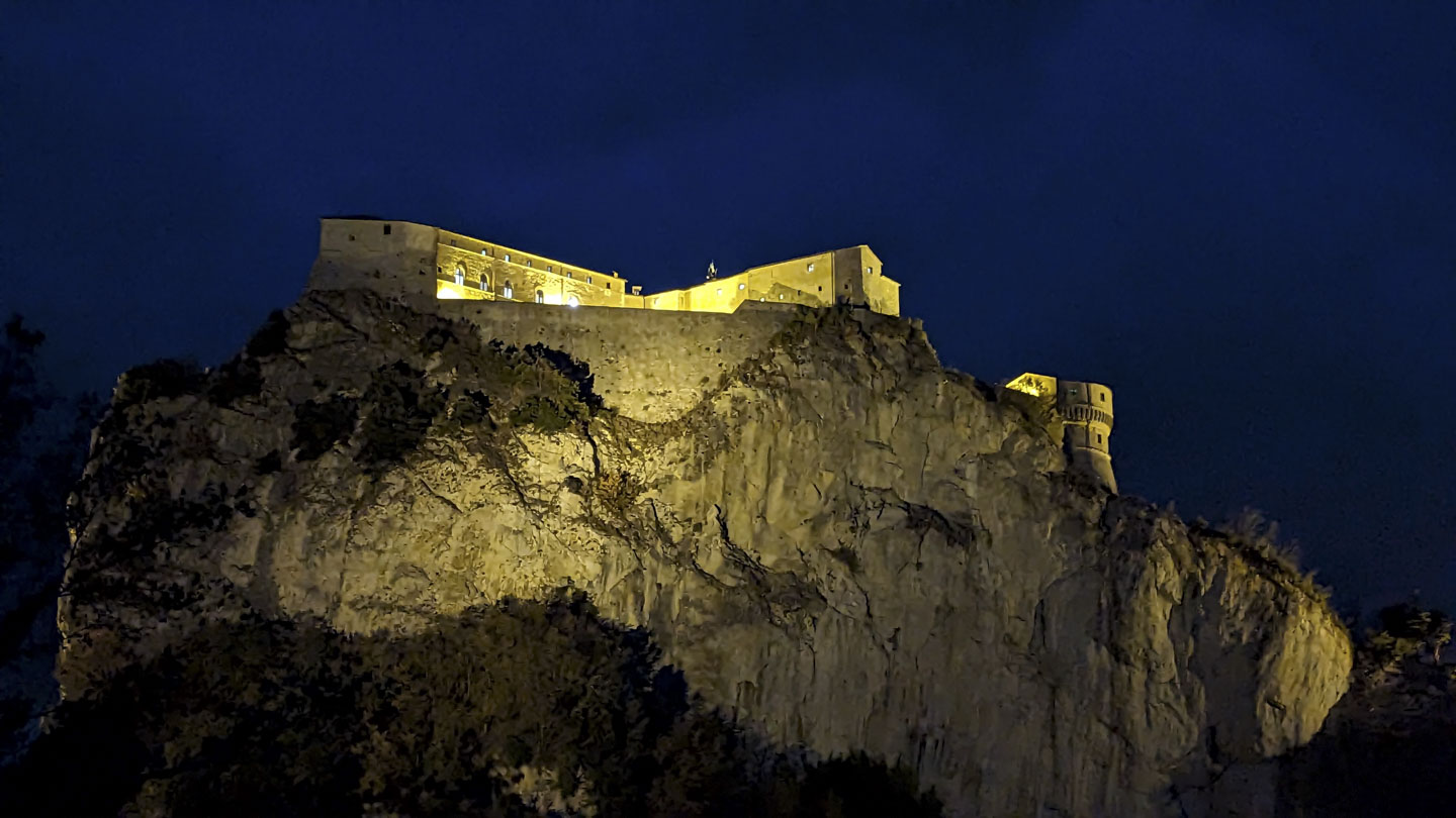 A sheer cliff wall with a stone fort on the top. Some soft yellow lights shine on the wall and the fort.