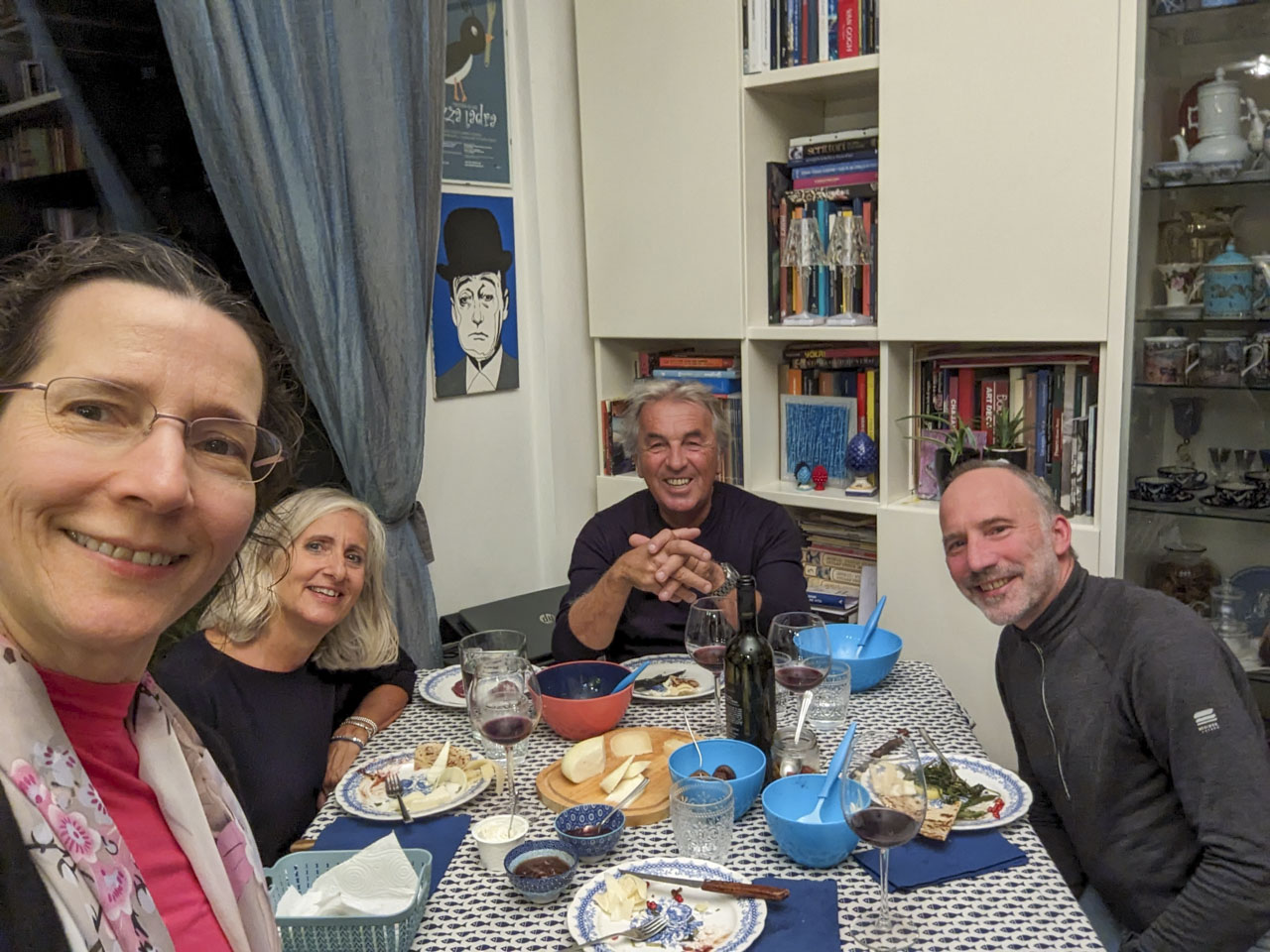 Alizia, Roberto, and Paul sit at a square table with remains of food in front of them. Anne is standing and taking a selfie.