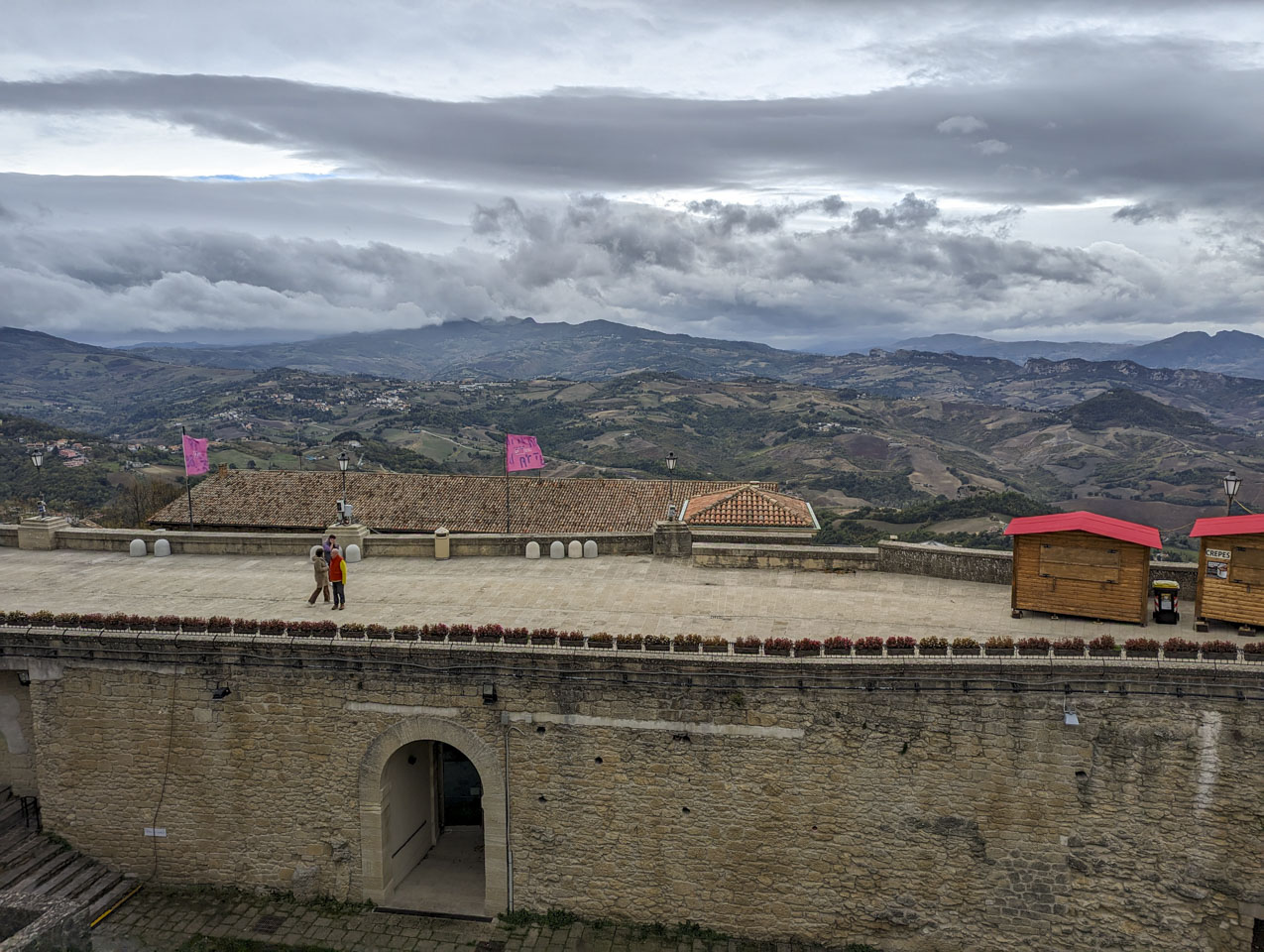 Part of a building is in the foreground, with a flat top that is a walking area. There are a few people on the area, and some closed vendor sheds. The land behind the building, which is much lower, has no flat areas. There are hills in the foreground and taller mountains in the background.