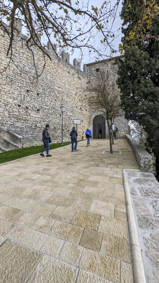 A tall stone wall is on the left, while 3 people walk up a stone walkway towards a door.