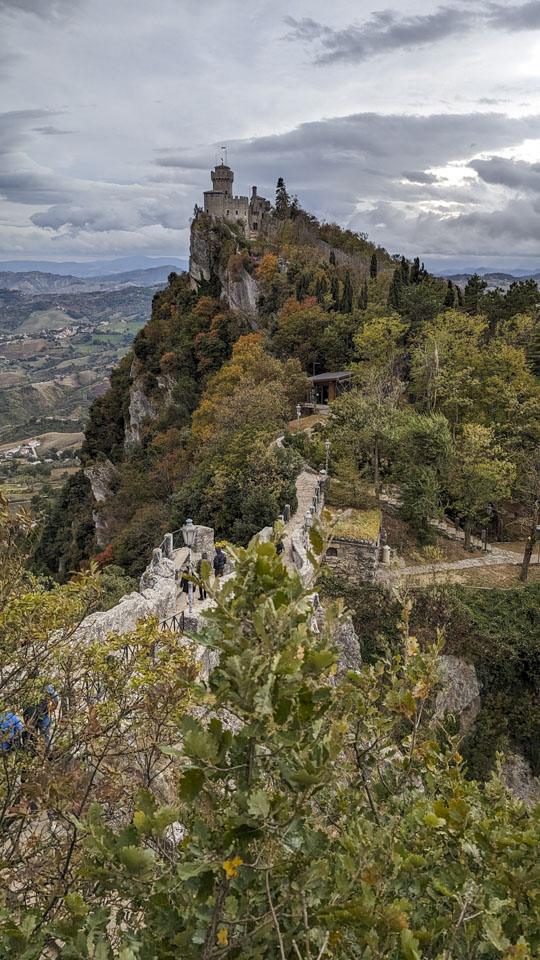 Looking down at a narrow path with a few people on it, and to a rising mountain top that has a tower on it. The land slopes off sharply to both sides.