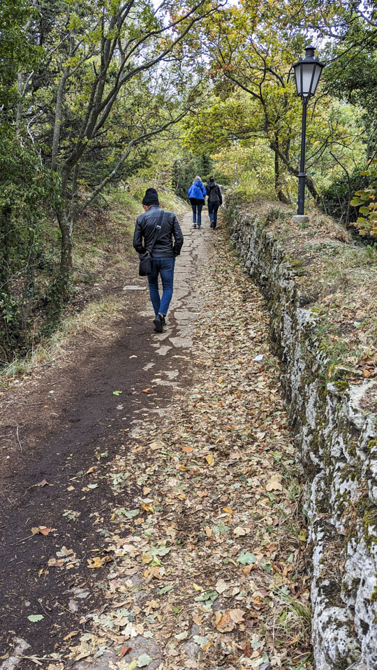 A path with some leaves, greenery on the left and a stone wall on the right. Roberto is walking away from us, then further away are Alizia and Anne.