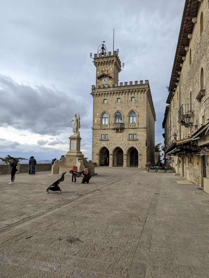 A plaza with a few people, a white statue, and a stone building at the far side.