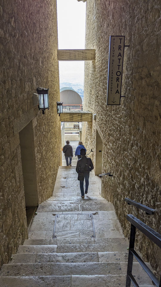 Looking down a long staircase with a couple of landings and tall stone building walls on each side. Anne and Alizia are side by side walking away in the distance and Roberto is behind them.