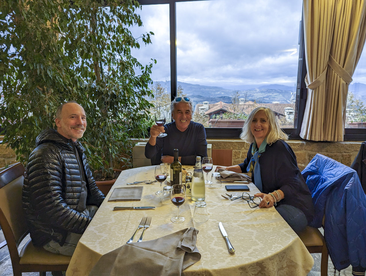 Paul, Robert, and Alizia at a table with a gold tablecloth. Roberto is holding up a glass of wine. Through the window mountains can be seen in the distance.