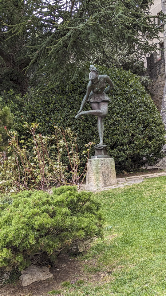 A statue of a ballet dancer standing on one leg while adjusting her footwear. The statue is in front of a green bush and has a bit of lawn in front of it.