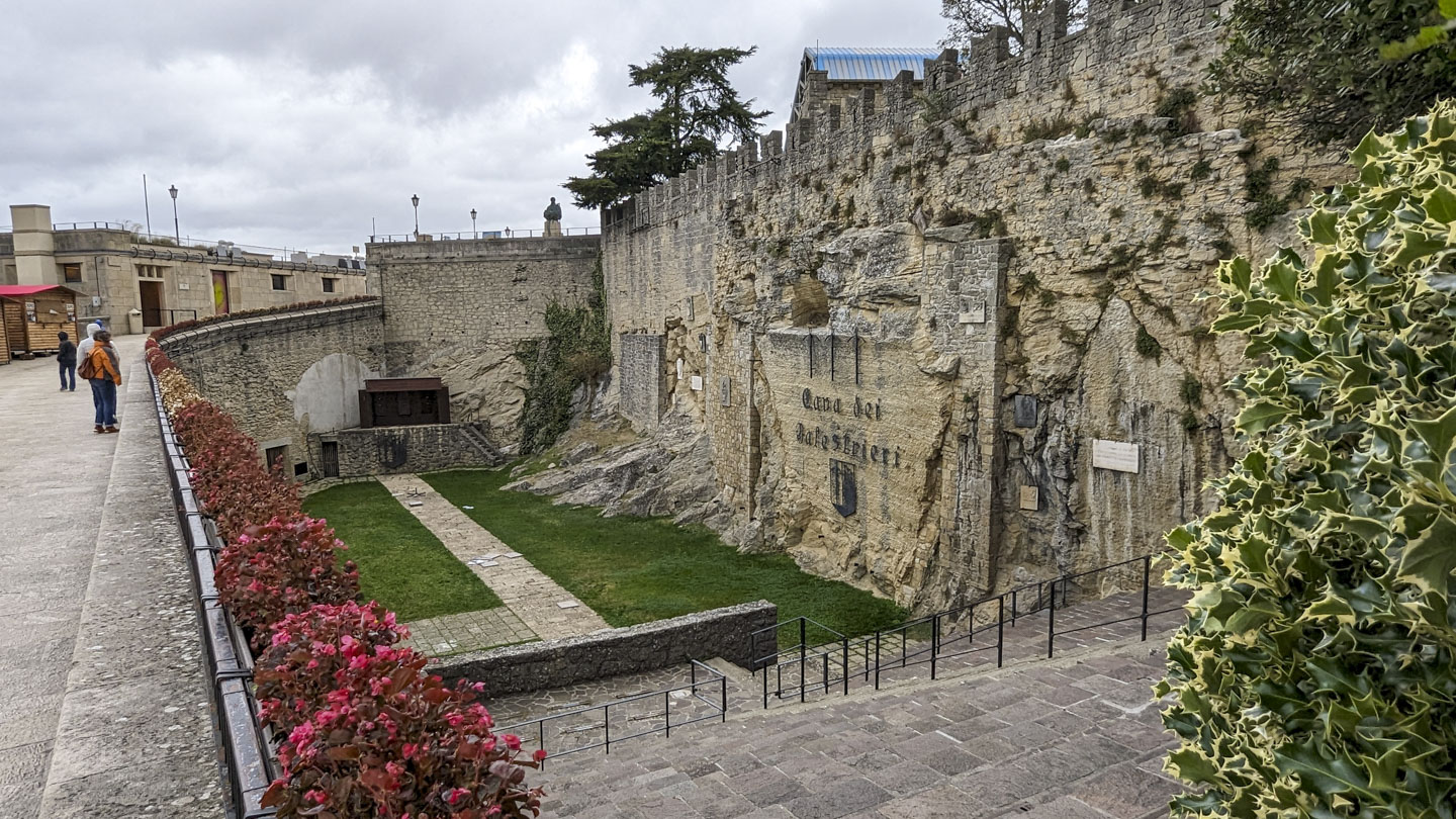 On the left of a photo the walkway continues level, but the rest of the photograph shows steps down to a long narrow sunken area that has green grass and stone walkways. At the far side is a raised platform of stone.