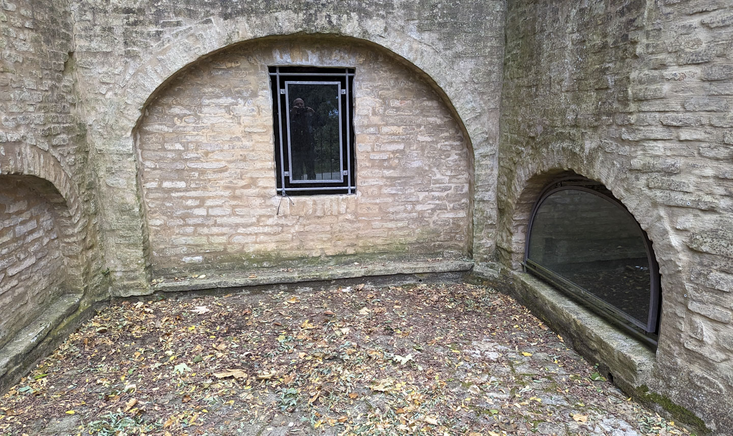 Paul reflection can be seen in some glass that covers an opening in the stone wall of a u-shaped area. There is also a window on the left, low to the ground.