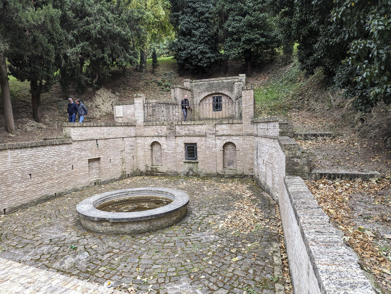 A u-shaped stone enclosure with a circular pool of water in the middle. It is set into a hillside. Two men are walking on the far side, and Paul is standing on a second level behind an iron picket fence.