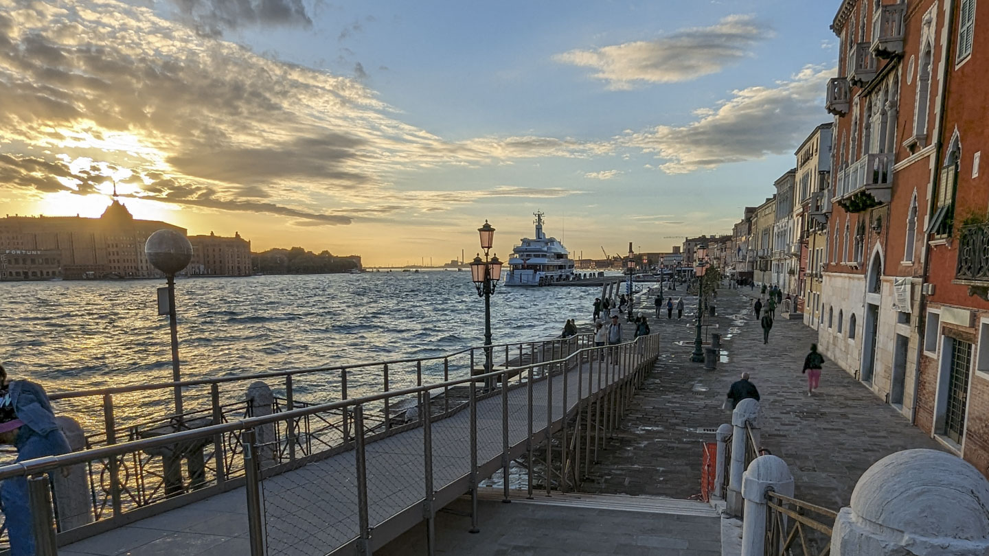 Taken from a bridge that also has a ramp, looking at the Grand Canal of Venice, with a large yacht in the water and the sun low in the sky in the left of the photo, behind some buildings.