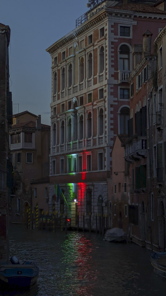 A canal in Venice in the evening, with red, green, and white lights shining on a house.
