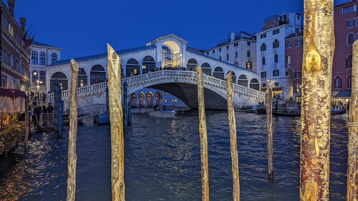 The Rialto bridge in the evening. In the foreground are piling that can be used by boats, and there is a boat going underneath the bridge.