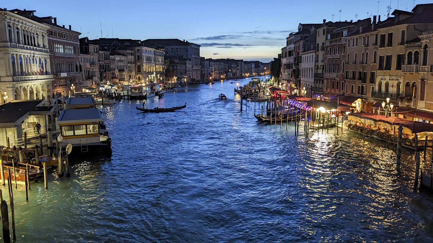 View from the Rialto Bridge in Venice. A waterbus stop is on the left, a gondola is crossing the canal towards boat slips on the right, with buildings and restaurants lining both sides of the canal.